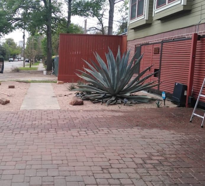 A restored brick paver driveway with sculptural agave planting against a corrugated red facade.
